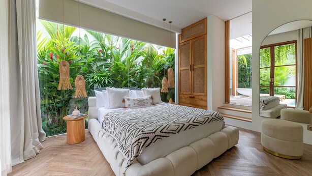 A bedroom with a large bed, a wooden side table, a mirror, and a view of lush greenery through large windows. - Villa BUMI - Designer Villa 10 min to the Beach (Bali)