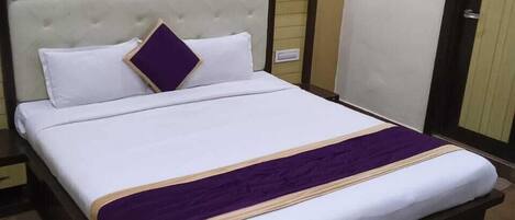 A neatly made bed with white bedding and a purple accent, a wooden headboard, and a wooden nightstand with a drawer.