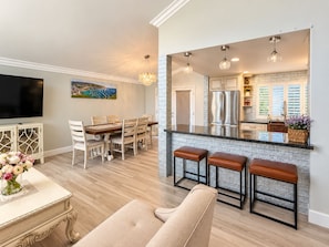 A modern kitchen with a central island, stainless steel appliances, and a dining area with a large TV and decorative flowers.