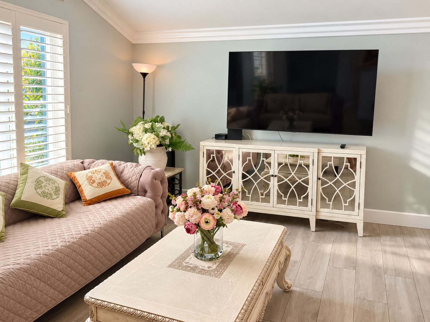 Un salon avec un canapé, une table basse ornée d’un vase de fleurs, un téléviseur posé sur un meuble et une fenêtre avec des stores.