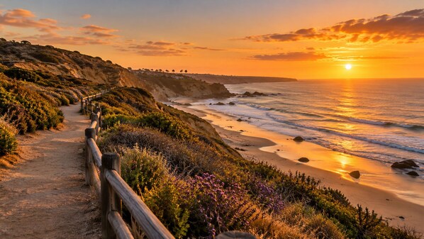 A coastal path with a wooden fence, leading to a beach with waves and a sunset.