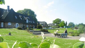Een woonwijk met een groot, wit gevelsteen huis, voorzien van een steile kap en meerdere ramen. Een verhard pad leidt naar een zitgedeelte met een tafel en stoelen.