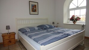 Een slaapkamer met een wit bed, een nachtkastje, een lamp, een raam met uitzicht en een lijst met foto aan de muur.