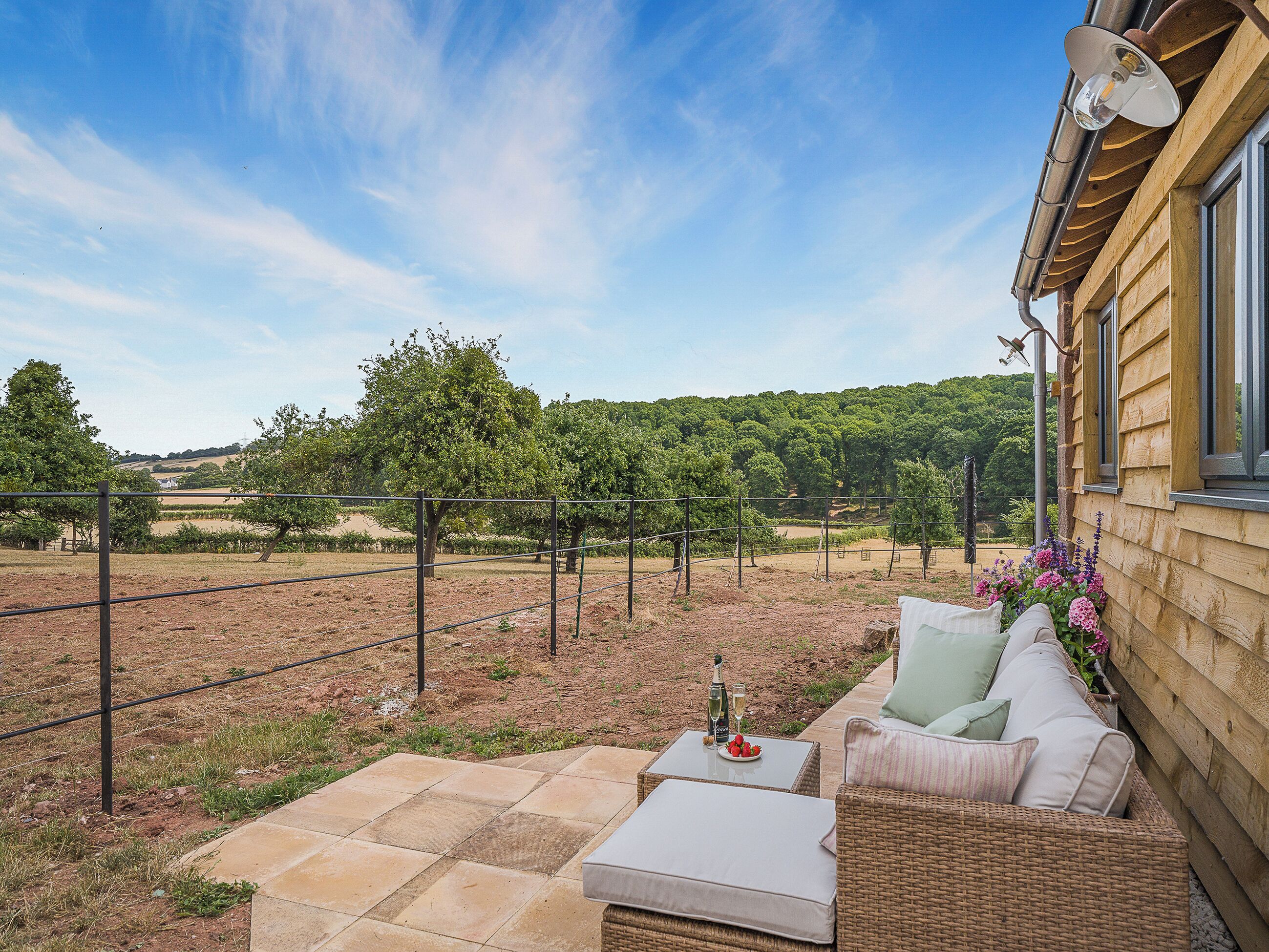 A patio with outdoor furniture, a table with a bottle and glasses, and a view of a vineyard and hills.