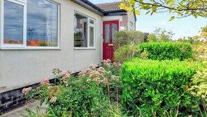 A residential house with a red door, large windows, and a garden with various plants and flowers. - 4 bedroom accommodation in Thornton Watlass, near Masham (Thornton Watlass, near Masham)