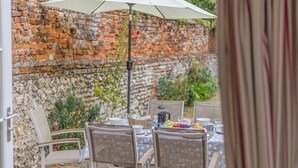An outdoor dining area with a brick wall, a white umbrella, wicker chairs, and a table set with plates, glasses, and a basket of bread.