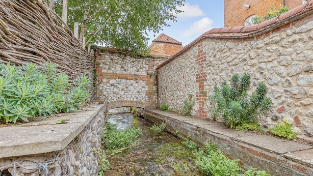 A narrow stone-lined water channel with vegetation on either side, flanked by stone walls and a wooden fence.