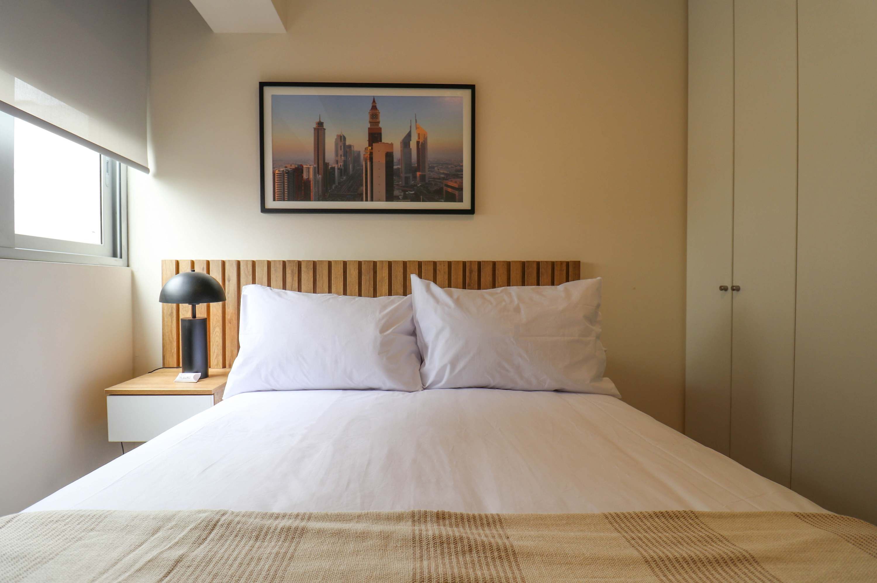 A hotel room with a bed, a nightstand, a lamp, a framed cityscape picture, and a wardrobe.