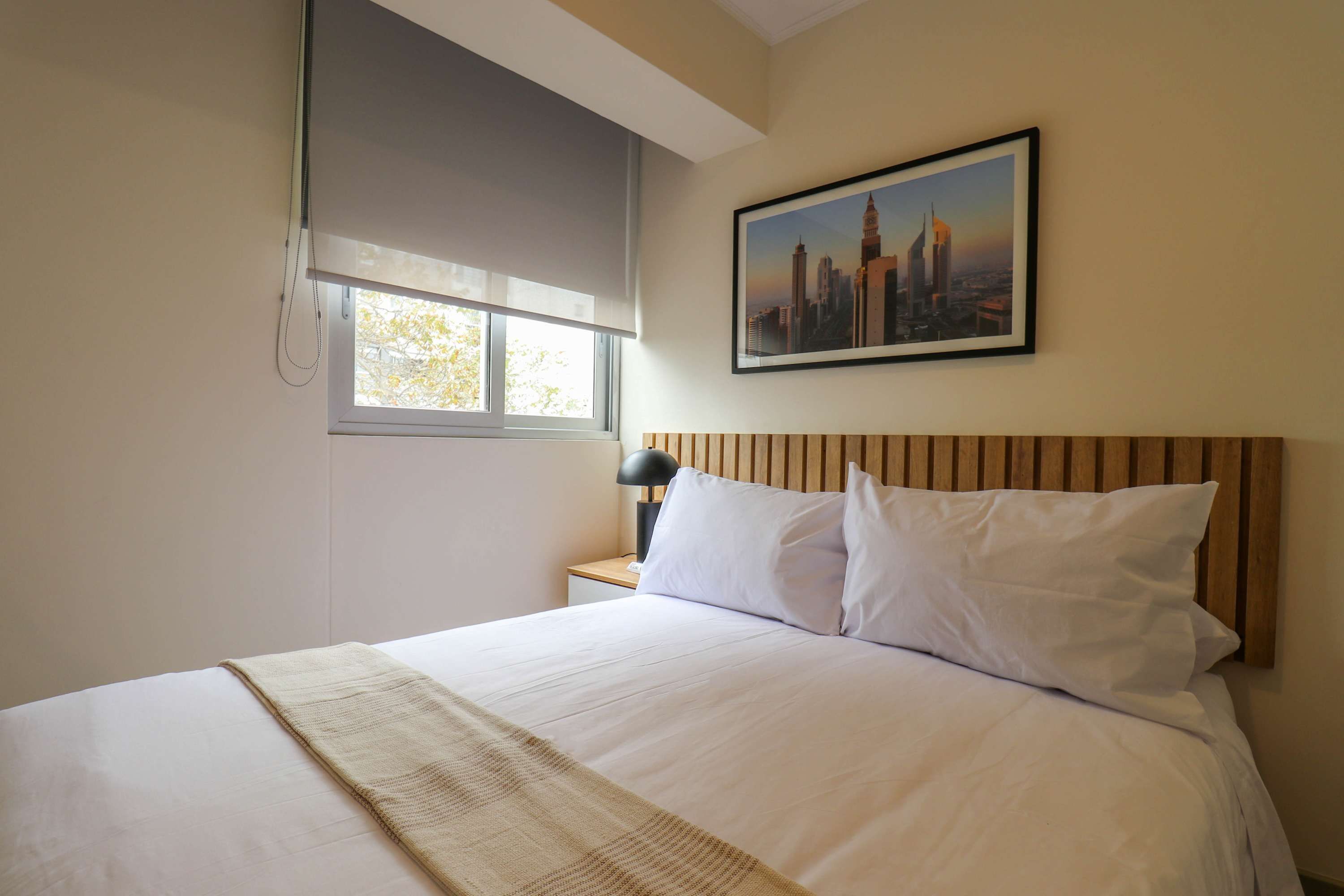 A hotel room with a bed, a nightstand, a lamp, a window with blinds, and a cityscape picture on the wall.