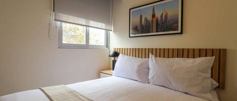 A hotel room with a bed, a nightstand, a lamp, a window with blinds, and a cityscape picture on the wall.