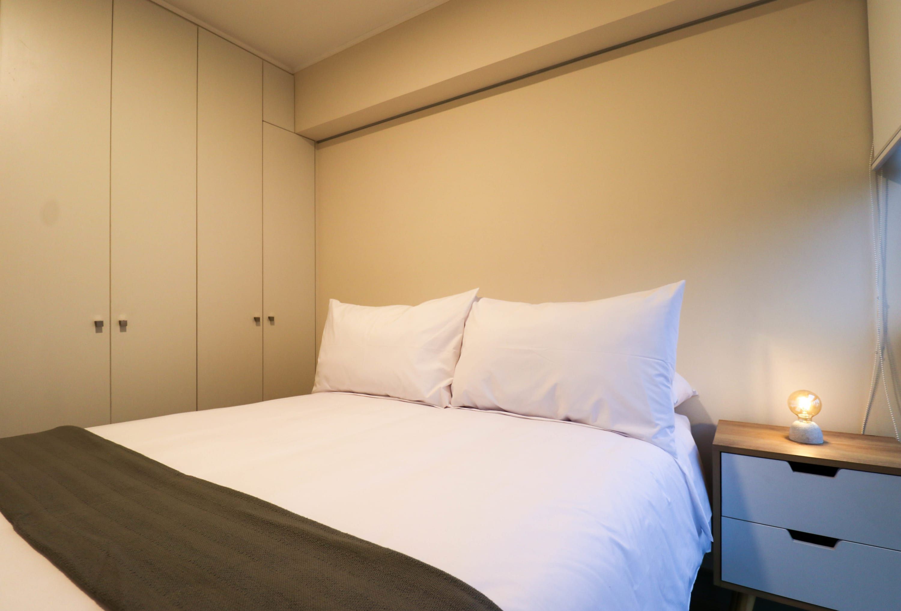 Una cama bien hecha con sábanas blancas y un edredón oscuro, una lámpara de noche y una mesita de noche de madera con cajón.