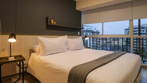 A modern hotel room with a large bed, a bedside table with a lamp, a small shelf with decorative items, and a view of the city through large windows. - w Glorious 2BR w Balcony in San Isidro (Lima)
