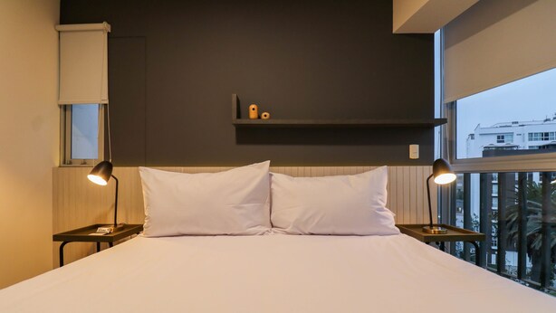A neatly made bed with white linens, two bedside lamps, a small shelf above the bed, and a window with a view of buildings. - w Glorious 2BR w Balcony in San Isidro (Lima)