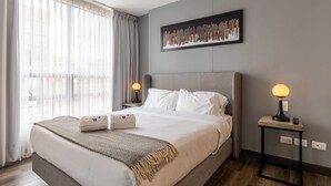 Ein modernes Hotelzimmer mit einem großen Bett, zwei Nachttischlampen, einem Wandbild und Blick durch leichte Vorhänge.