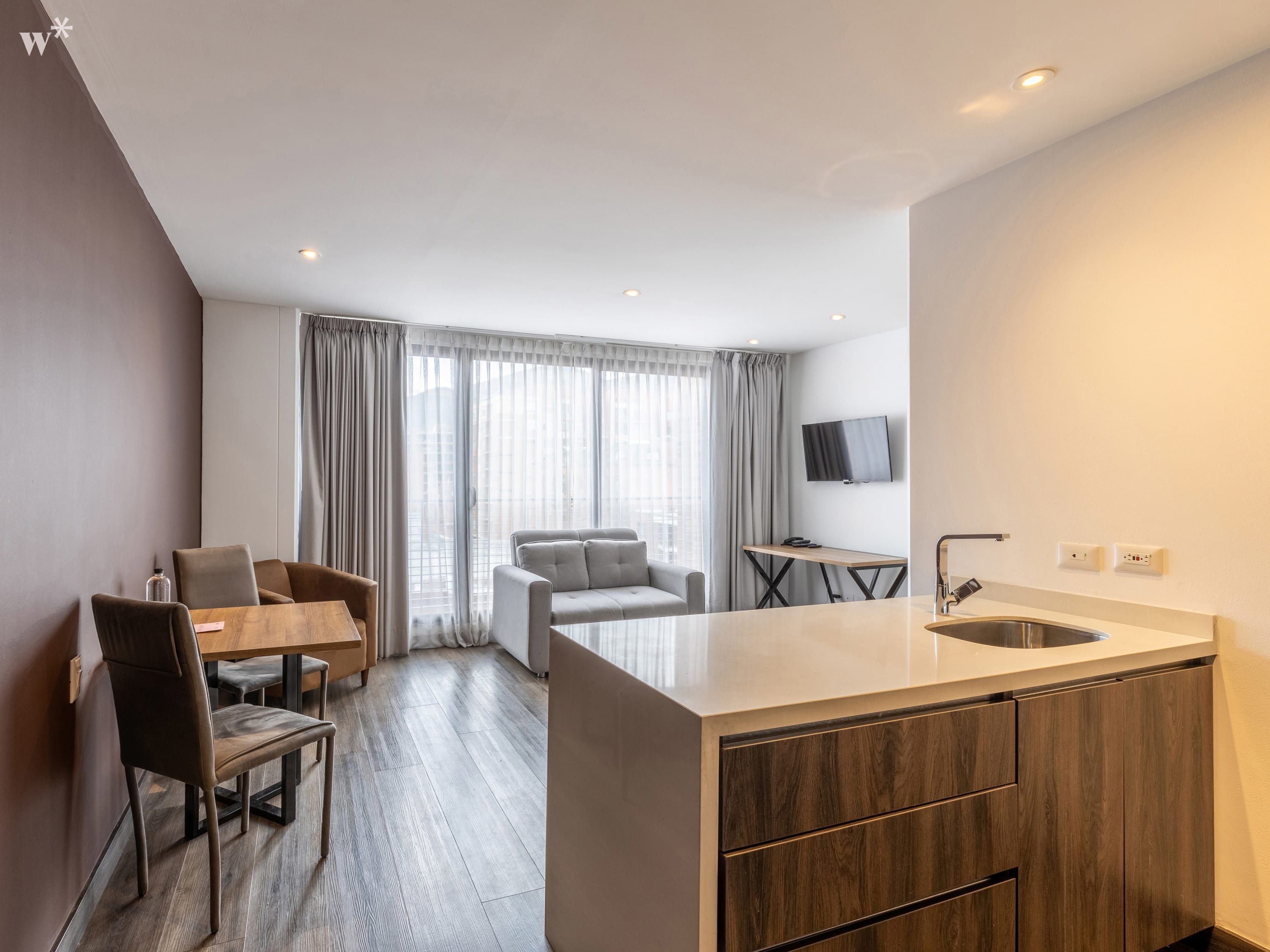 Een moderne keuken met een centraal eiland, een eethoek met tafel en stoelen, een bank en een aan de muur bevestigde televisie.