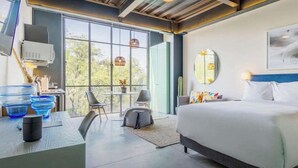 Habitación de hotel moderna con una cama grande, un escritorio con computadora, una silla y vistas a árboles a través de grandes ventanales.