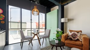 Una sala de estar moderna con puerta de vidrio que da a un balcón, un sofá marrón, dos sillas, una mesita con una planta y una lámpara colgante.