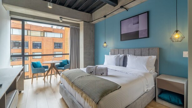 A modern bedroom with a large bed, a desk, and chairs, a view of a brick building, and a blue accent wall. - w Dreamy 2BR in Parque Virrey (Bogotá)