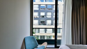 A room with a large window, a chair, a bed, and a view of a building outside.