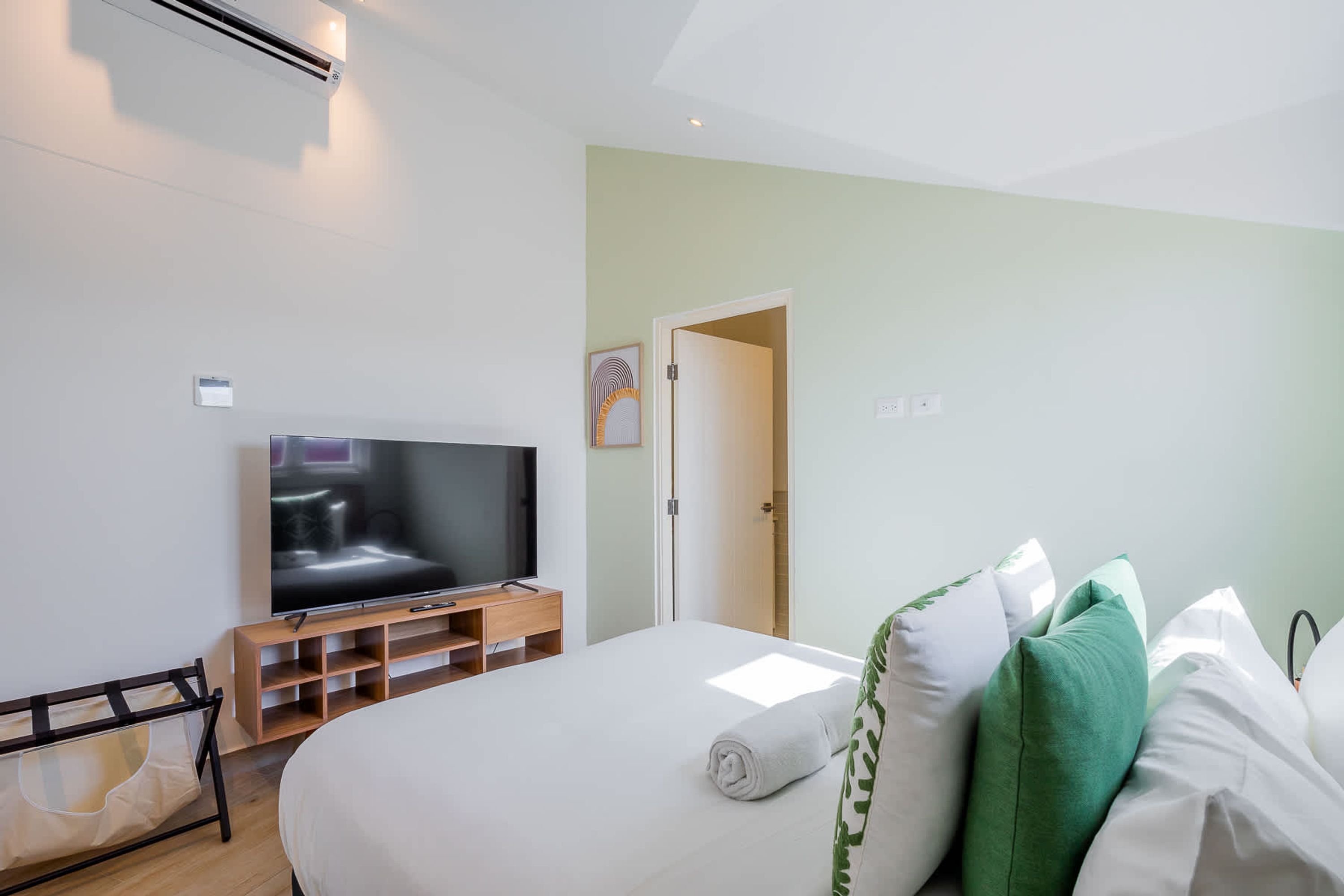 Hotellihuone, jossa on sänky, televisio, puinen yöpöytä ja ovi, joka johtaa toiseen huoneeseen.