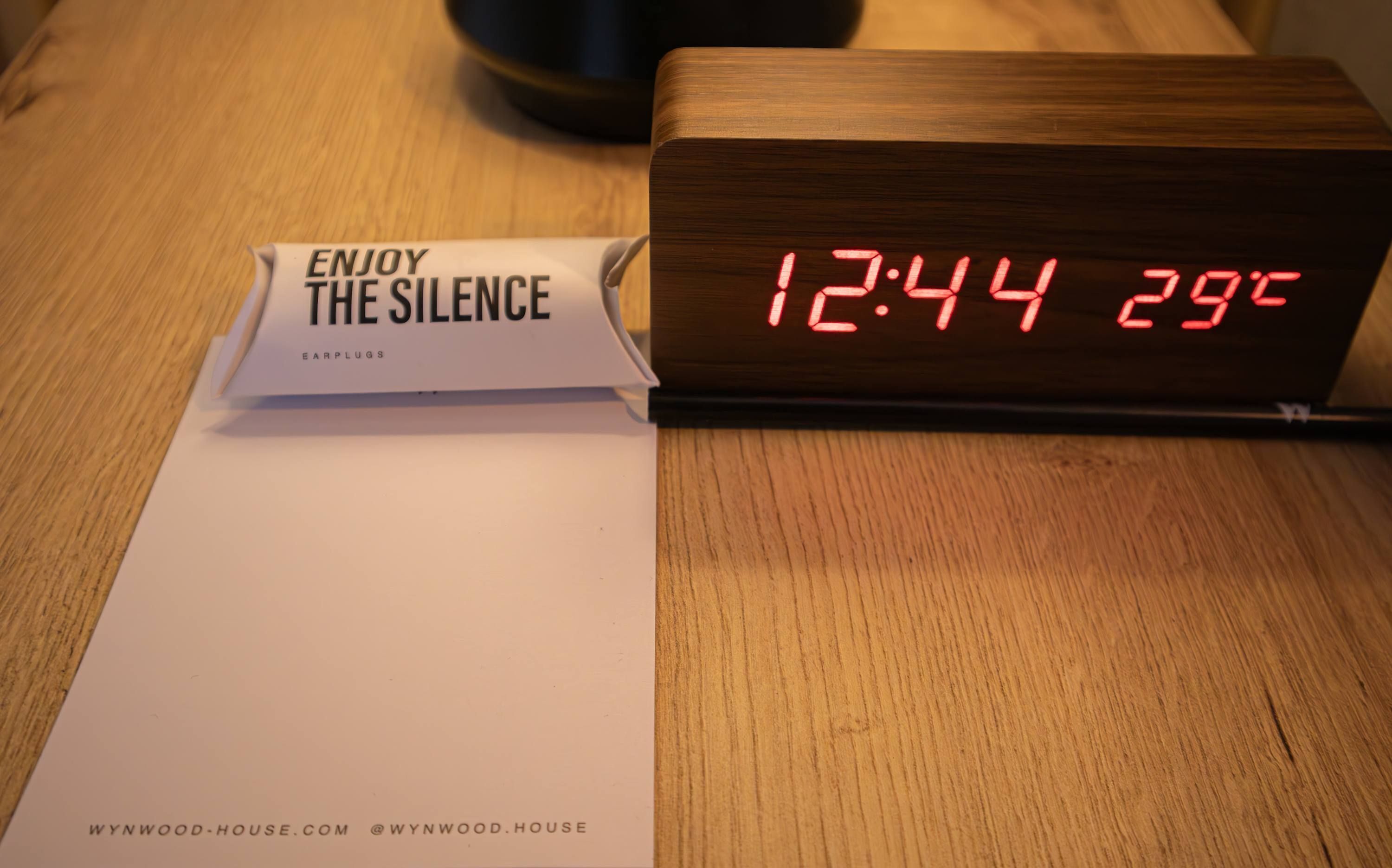 A wooden table with a digital clock displaying 12:44 and a sign that reads 'ENJOY THE SILENCE EARPLUGS.'