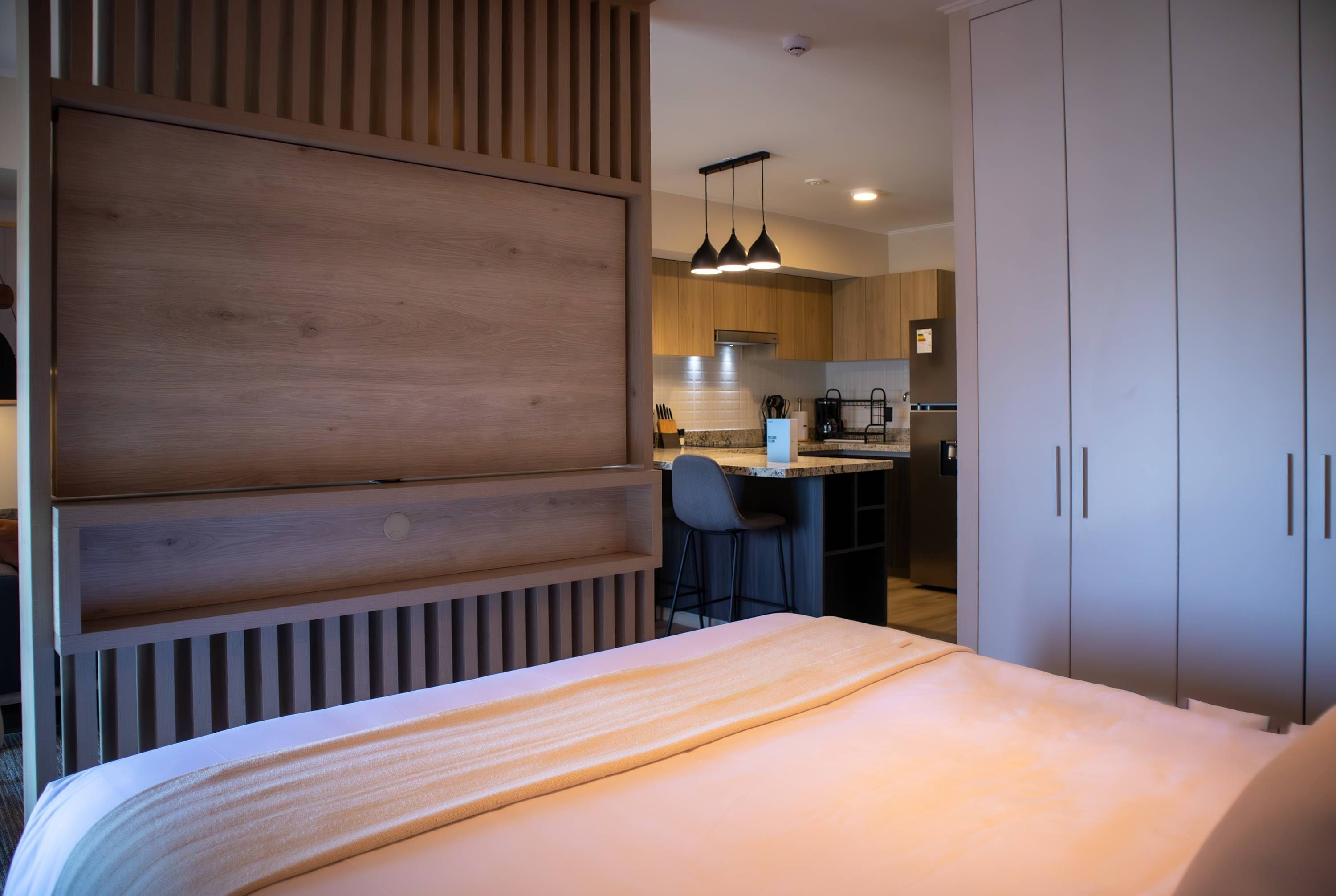A modern hotel room with a wooden panel wall, a bed with a striped blanket, a small kitchen area with a bar stool, and a dining table.