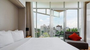 Une chambre d’hôtel moderne avec un grand lit, un canapé gris agrémenté d’un coussin rouge et une vue sur la ville grâce à de grandes fenêtres du sol au plafond.
