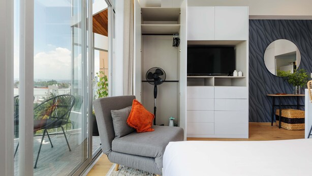 Une chambre moderne avec un canapé gris, une petite table, un téléviseur et une grande fenêtre donnant sur la ville.