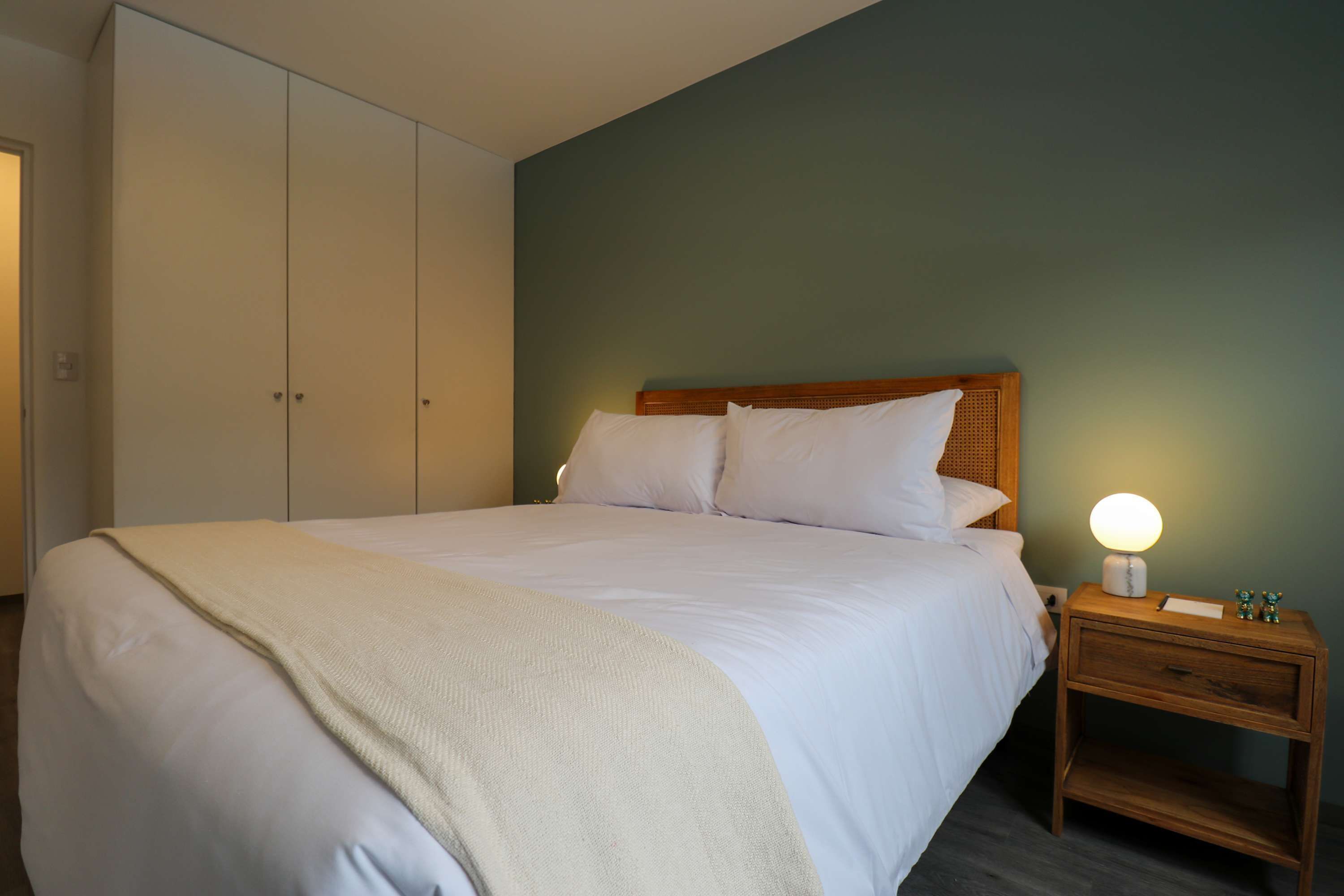 Una cama bien hecha con sábanas blancas y cabecera de madera en una habitación con paredes verde azulado y una mesita de noche de madera con lámpara.