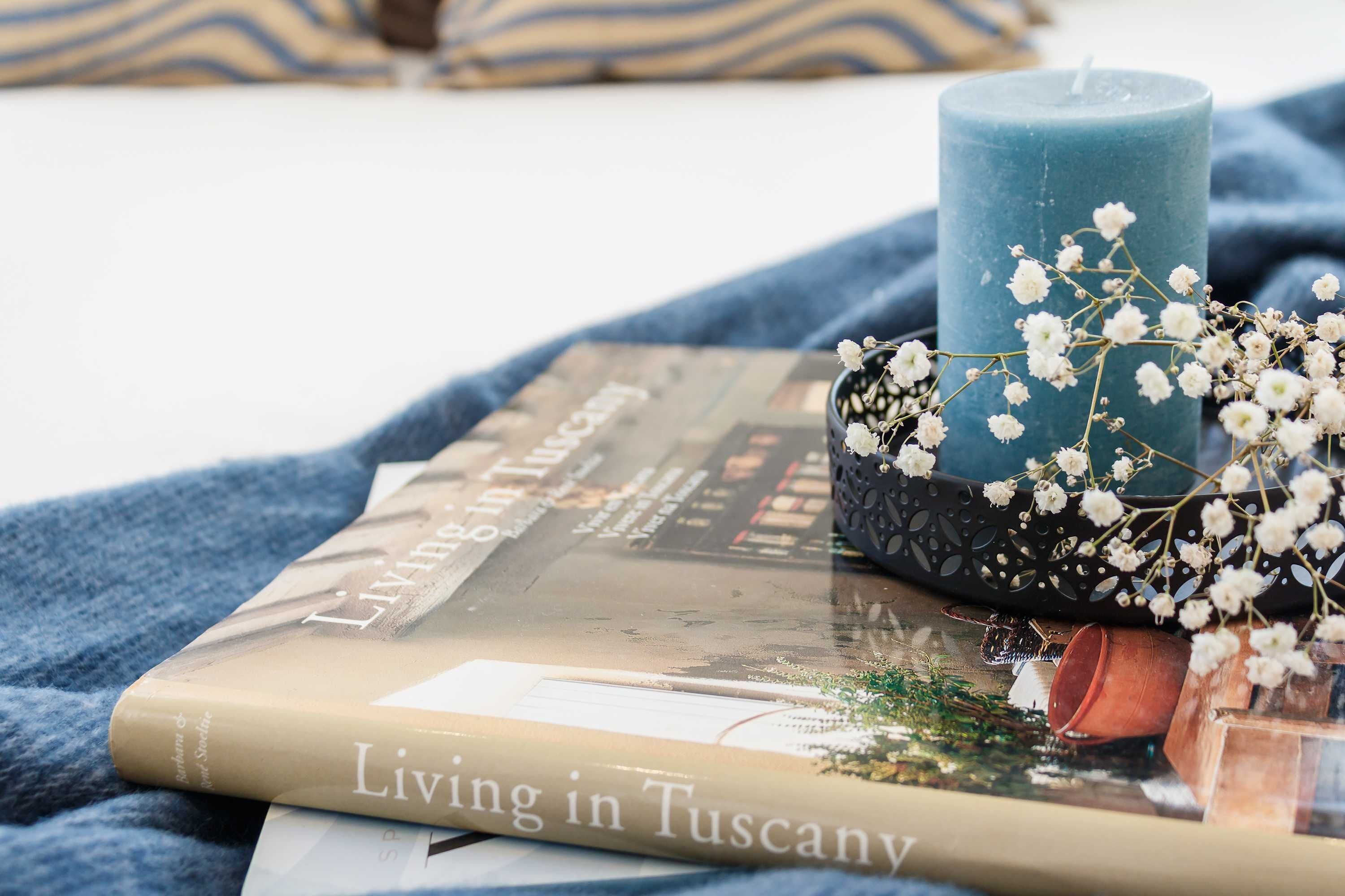 A book titled "Living in Tuscany" on a blue blanket with a candle and white flowers.