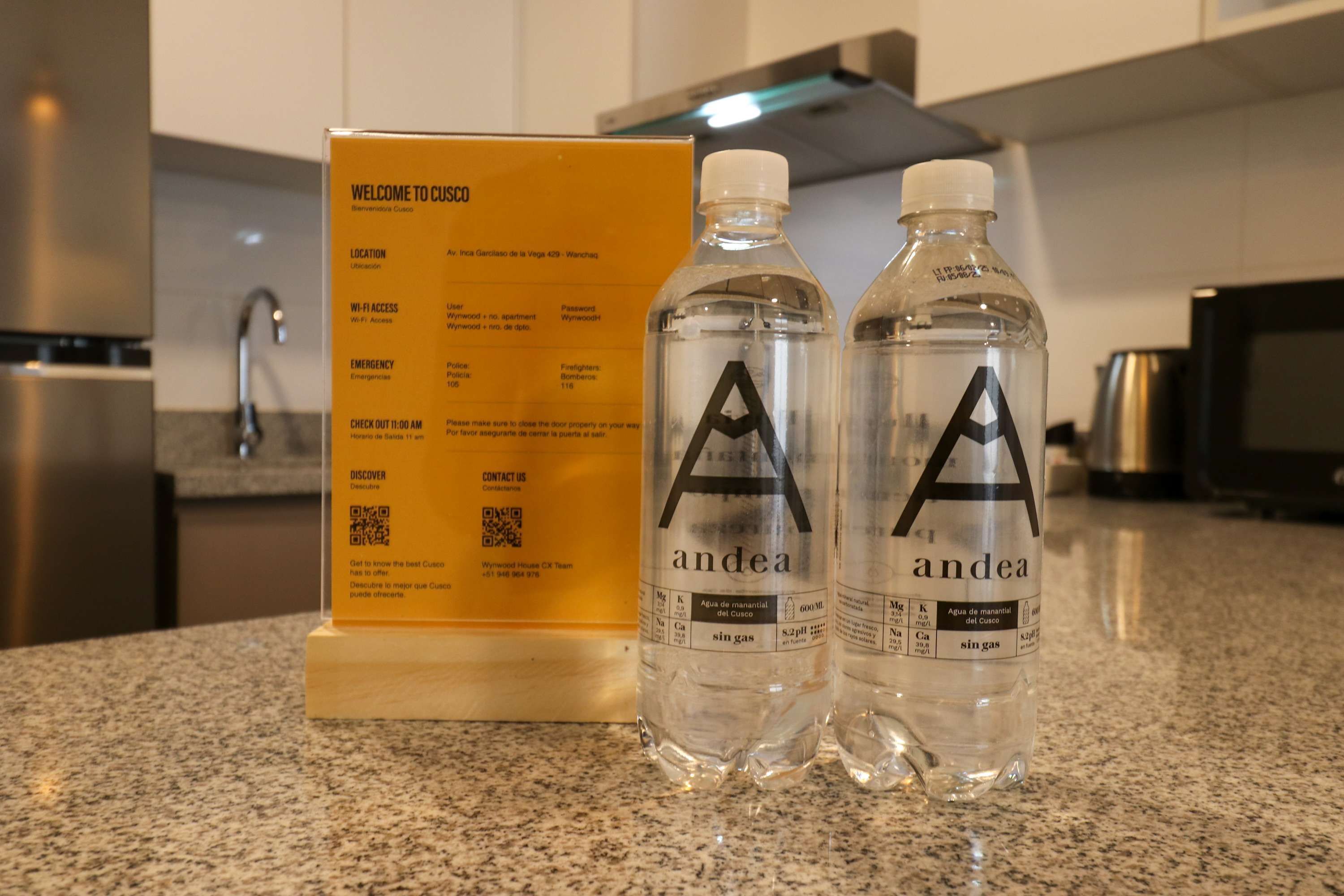 A welcome sign with location details, emergency contacts, and a QR code. Two water bottles with the brand 'andea' are on a kitchen counter.