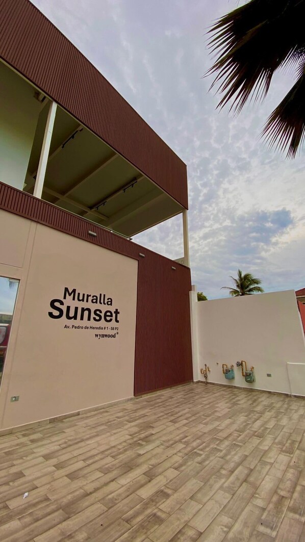A building named Muralla Sunset with a large sign on the facade, a wooden floor, and a palm tree in the background.