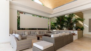 Amplia sala de estar con un sofá seccional grande, un sofá más pequeño y una chaise lounge. Hay una mesa de centro y una mesita auxiliar. La habitación tiene un techo alto con diseño de listones de madera y ventanas grandes que permiten la entrada de luz natural.