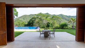 Una puerta de madera abierta que da a una terraza con mesa y sillas, con vistas a una piscina y abundante vegetación, con montañas al fondo.
