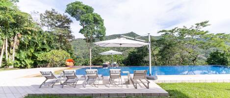 A poolside area with lounge chairs and umbrellas, surrounded by lush greenery and a mountainous backdrop.