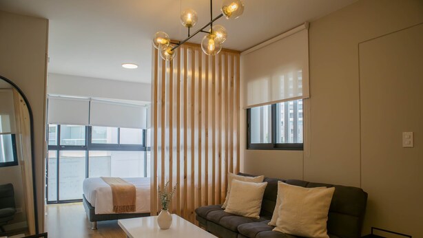 A modern living room with a sofa, a coffee table, and a bed. There is a large window with blinds, a wooden slat partition, and a hanging light fixture. - w Stunning 1BR w Great Deco in San Isidro (Lima)
