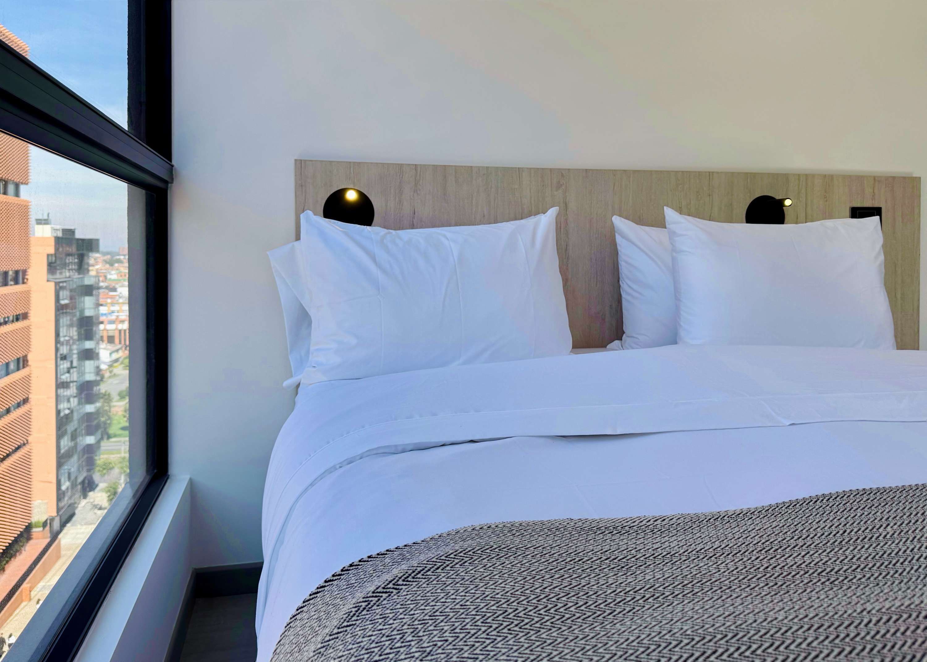 A hotel room with a large bed, a view of the city through a window, and a modern headboard with circular light fixtures.