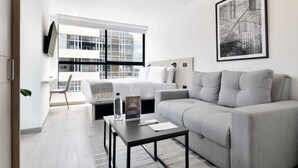 Una habitación de hotel moderna con un sofá gris, una mesita con una botella y un libro, una cama con ropa de cama blanca y un ventanal con vistas a la ciudad.