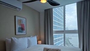 A hotel room with a large bed, a ceiling fan, a framed abstract painting, a bedside lamp, and a view of a cityscape through a large window. - w Awesome 1BR w Great Deco in Avenida Balboa (Panama City)