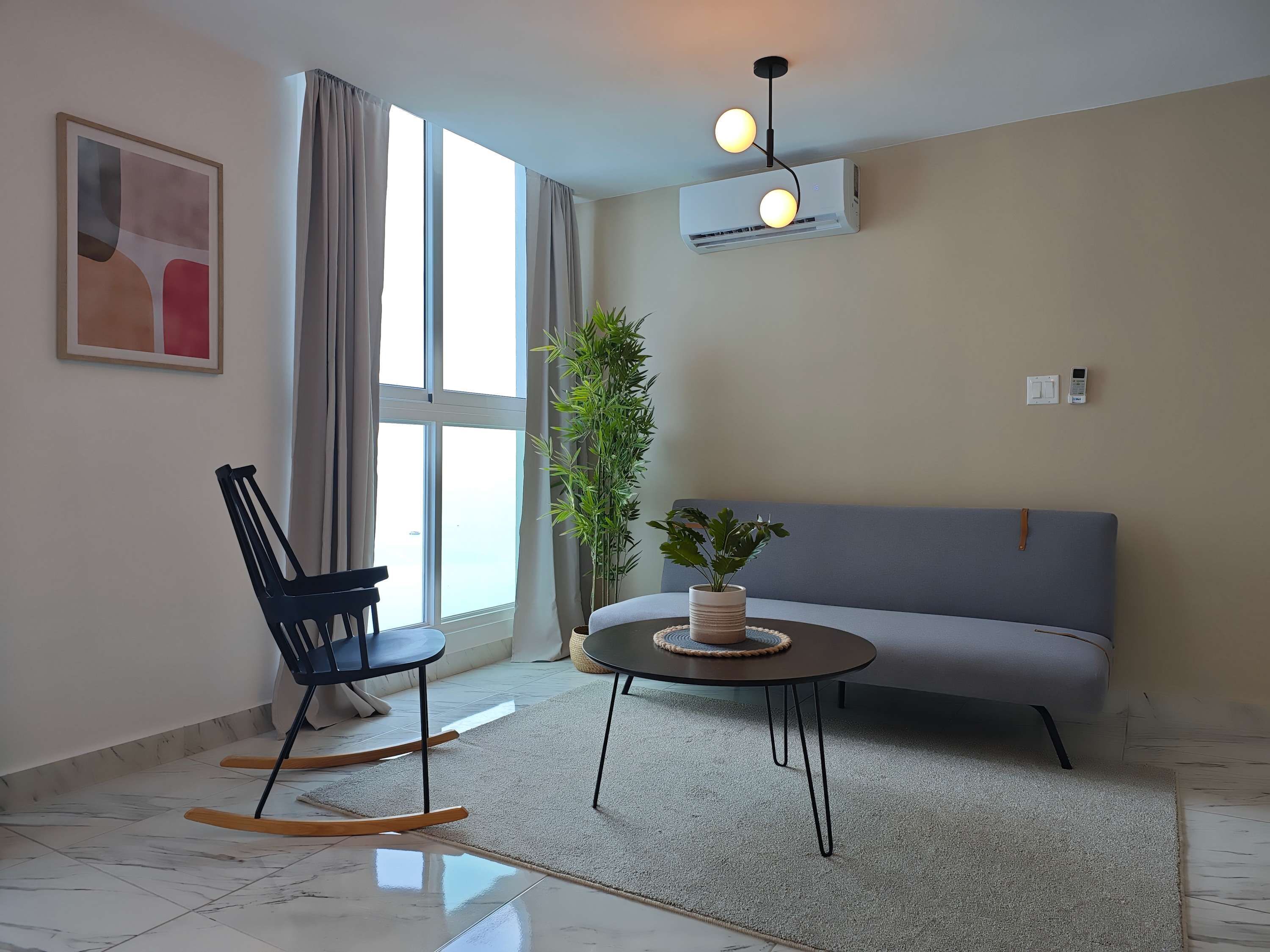 Una sala de estar moderna con sofá, sillón mecedora, mesa de centro, planta en maceta y equipo de aire acondicionado.