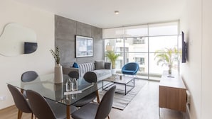 A modern living room with a glass dining table, grey sofa, blue armchair, and a TV mounted on the wall. - w Great 3BR Duplex With Terrace in Miraflores (Lima)