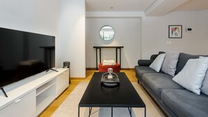 Uma sala de estar moderna com uma televisão de tela plana, uma mesa de centro preta, um sofá cinza e um banquinho vermelho.