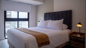 Une chambre d’hôtel moderne avec un grand lit, une fenêtre donnant sur des immeubles, une table de chevet avec une lampe et une tête de lit en bois.