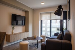 Een moderne woonkamer met een bank, een ronde salontafel, een aan de muur bevestigde televisie en een groot raam met jaloezieën.