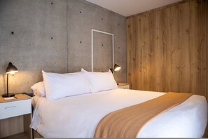 A neatly made bed with white linens and a brown throw blanket, flanked by two bedside tables with lamps, in a room with a concrete and wooden wall paneling. - w Phenomenal 1BR w Balcony in Barranco (Lima)