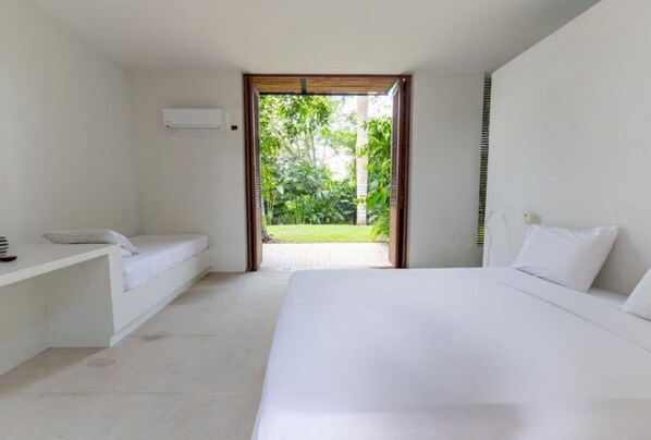 Une chambre à coucher au style minimaliste, avec un grand lit, une table de chevet et une vue sur un jardin luxuriant, visible à travers une porte ouverte.