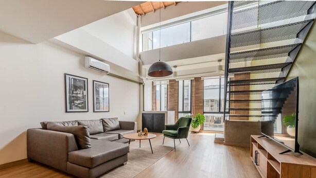 A modern living room with a sofa, a coffee table, and a TV stand. There is a staircase with a railing and a plant on the floor. - w Stylish Duplex in Laureles (Medellín)