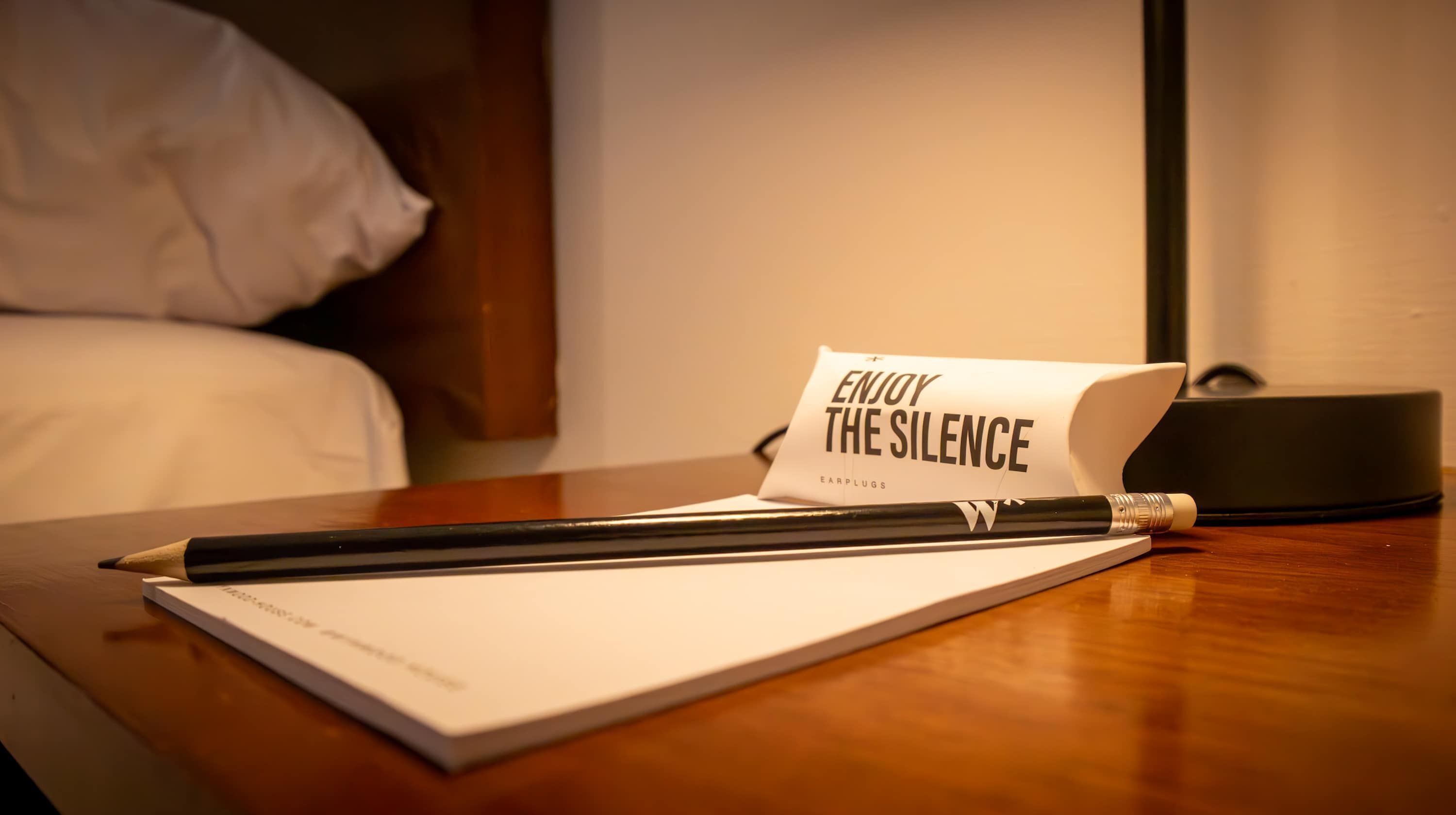 Ein Hotelzimmer mit einem Bett, einem Nachttisch, einem Buch mit dem Titel 'Enjoy the Silence', einem Bleistift und einem schwarzen Gegenstand, vermutlich einer Fernbedienung.