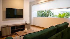 A modern living room with a flat-screen TV mounted on the wall, a wooden entertainment unit, a green sofa, a small table with a vase, and a large window with a view of greenery. - w Striking 1BR in Miraflores (Lima)