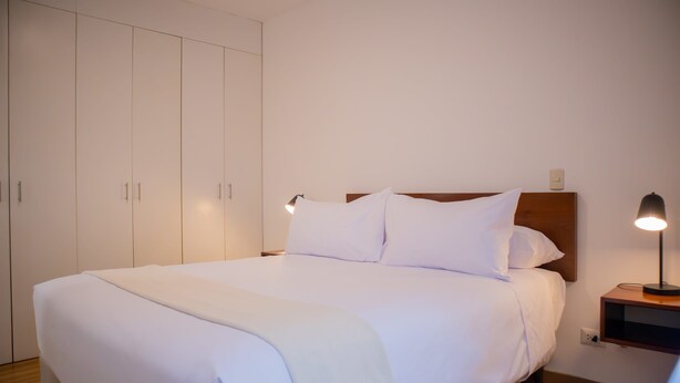 A neatly made bed with white linens and a wooden headboard, a bedside table with a lamp, and a wardrobe with sliding doors. - w Striking 1BR in Miraflores (Lima)